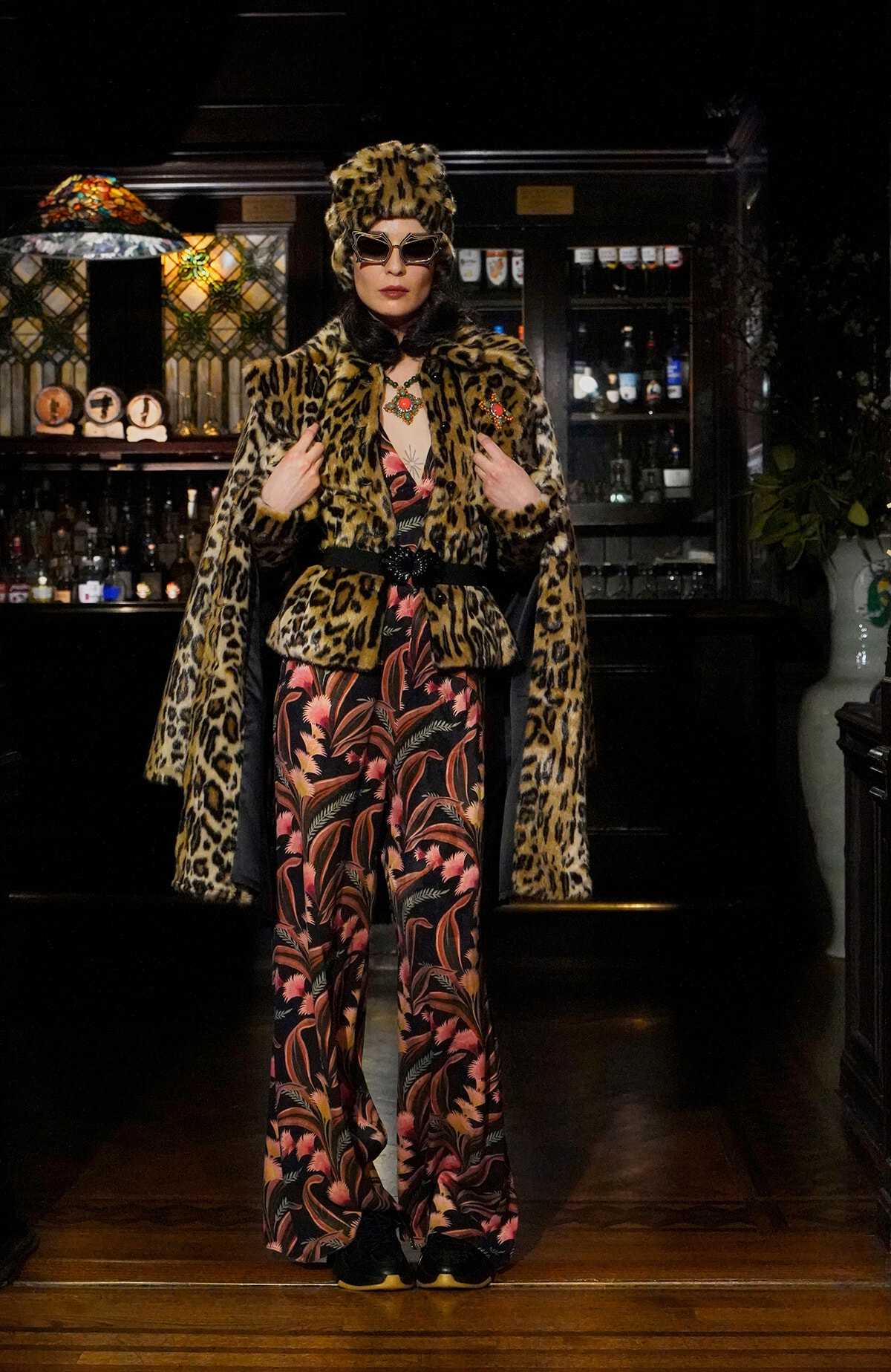 Person wearing a leopard print fur coat, matching hat and sunglasses posing in a bar setting with bottles in the background.