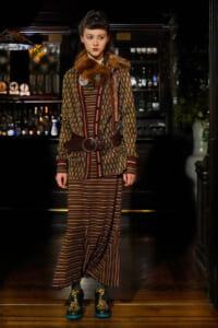 Model wearing a vintage geometric coat with a brown fur collar, cinched with a wide belt, over a striped dress in a bar-like setting.