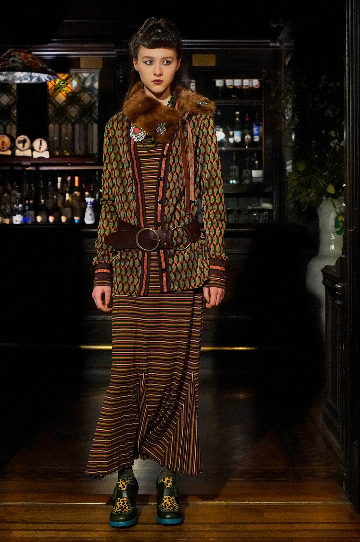Model wearing a vintage geometric coat with a brown fur collar, cinched with a wide belt, over a striped dress in a bar-like setting.