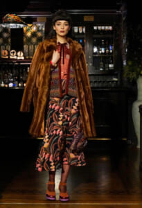 Model in a brown fur coat over a patterned dress, standing in a dim bar with bottles behind her.