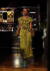 Model wearing an olive-green floral dress with large front bows, fur cuffs, and teal platform shoes in a dim bar setting.