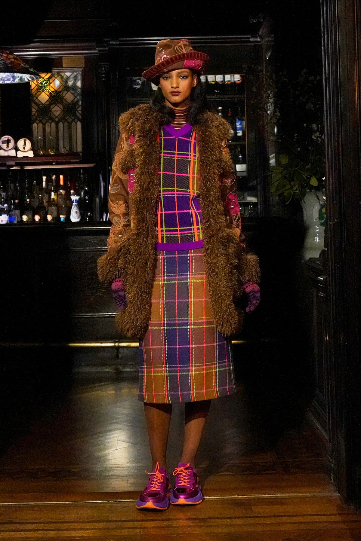 Person standing in a bar wearing a brown faux-fur coat over a bright plaid dress, pink hat, and purple sneakers.