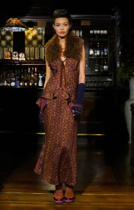Model wearing a tailored brown-purple patterned floor-length dress with a furry brown vest in a bar setting, accessorized with a red necklace.