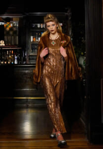 Model wearing a bronze metallic pleated jumpsuit with a brown fur coat, posing in a dimly lit bar setting.