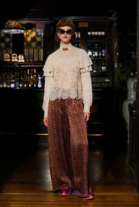 Model in a bar wearing a cream lace blouse with ruffles, brown patterned wide-leg pants, and cat-eye sunglasses.
