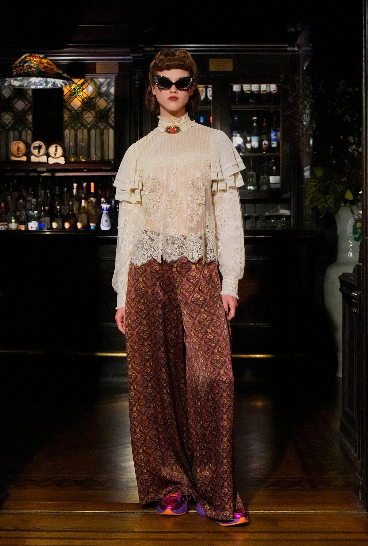 Model in a bar wearing a cream lace blouse with ruffles, brown patterned wide-leg pants, and cat-eye sunglasses.