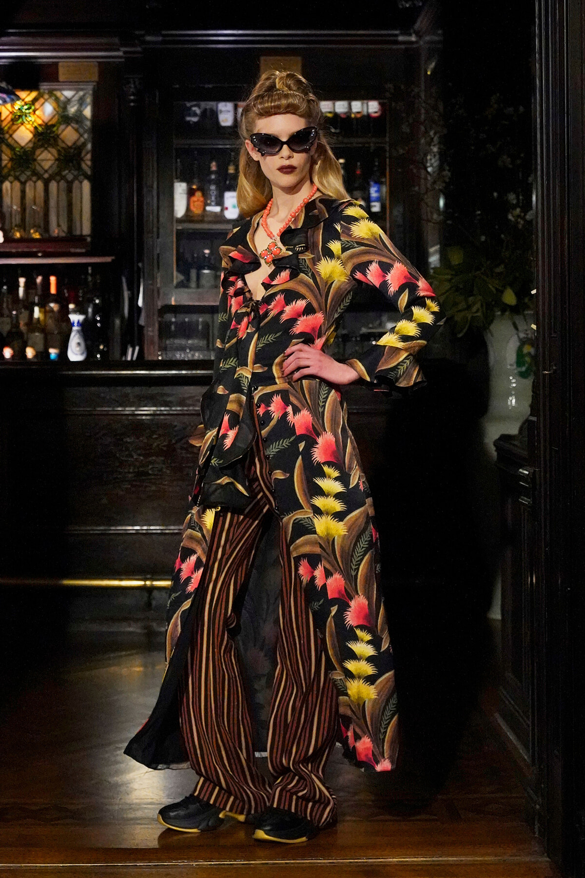 Model in a tropical-floral coat and striped pants posing in a dimly lit bar, wearing cat-eye sunglasses and a coral necklace.