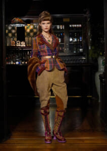Model in a colorful plaid blazer, wide belt, tweed pants, fur stole, and leopard boots posing in a dimly lit bar interior with bottles behind.