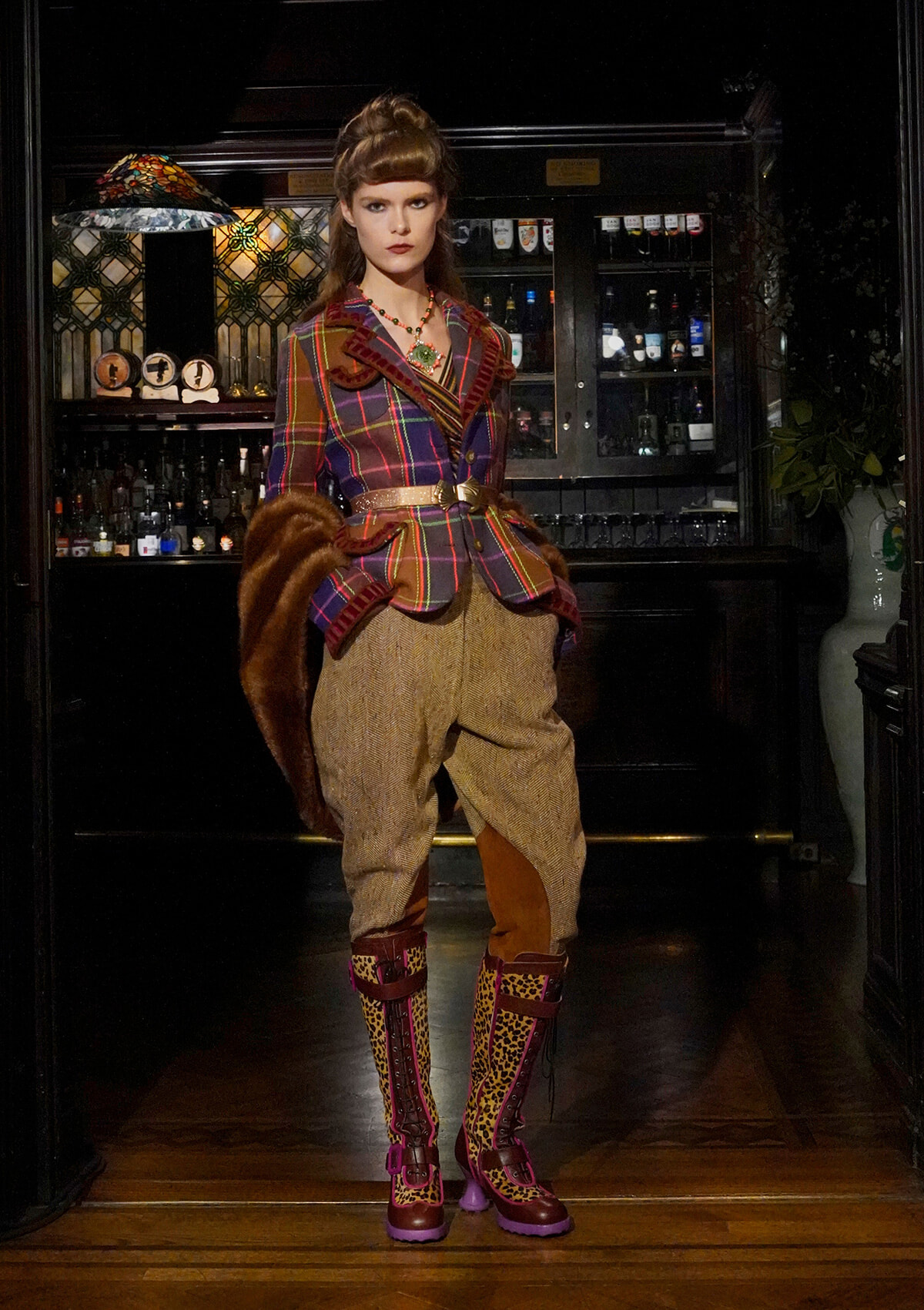 Model in a colorful plaid blazer, wide belt, tweed pants, fur stole, and leopard boots posing in a dimly lit bar interior with bottles behind.