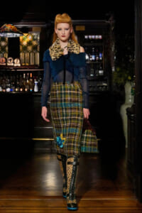 Model walks through a dim bar backdrop in a bold teal off-shoulder top with leopard fur collar, plaid midi skirt, leopard boots, and a blue handbag.