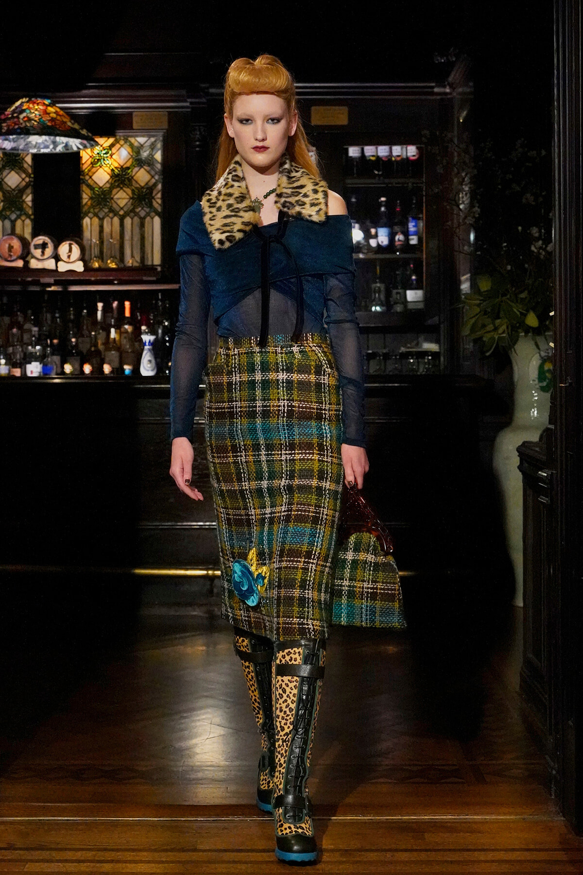 Model walks through a dim bar backdrop in a bold teal off-shoulder top with leopard fur collar, plaid midi skirt, leopard boots, and a blue handbag.