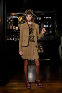 Model in a leopard-print outfit: camel tweed blazer and pleated skirt, leopard hat, matching bag, patterned tights, and pink leopard‑print shoes in a bar setting