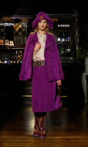 Model in a textured purple outfit: oversized coat, matching hat, beige blouse, wide belt, leopard-heeled boots, and a small purple handbag in a bar setting