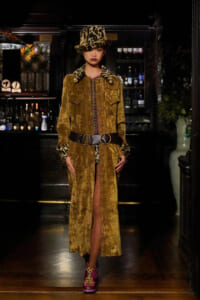 Model wearing a mustard velvet long coat with leopard fur cuffs and matching leopard-print hat, standing in a bar.