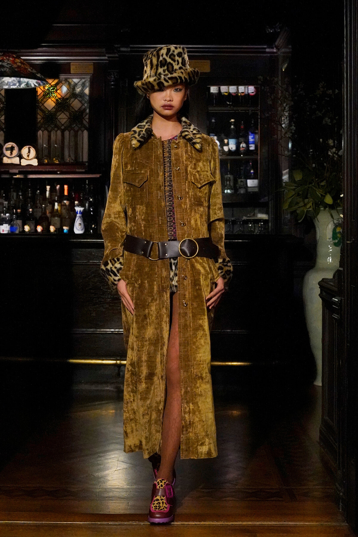 Model wearing a mustard velvet long coat with leopard fur cuffs and matching leopard-print hat, standing in a bar.
