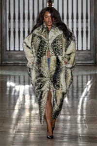 Model walks a fashion runway wearing an oversized leopard-print fur coat and black heels, with hands in coat pockets.