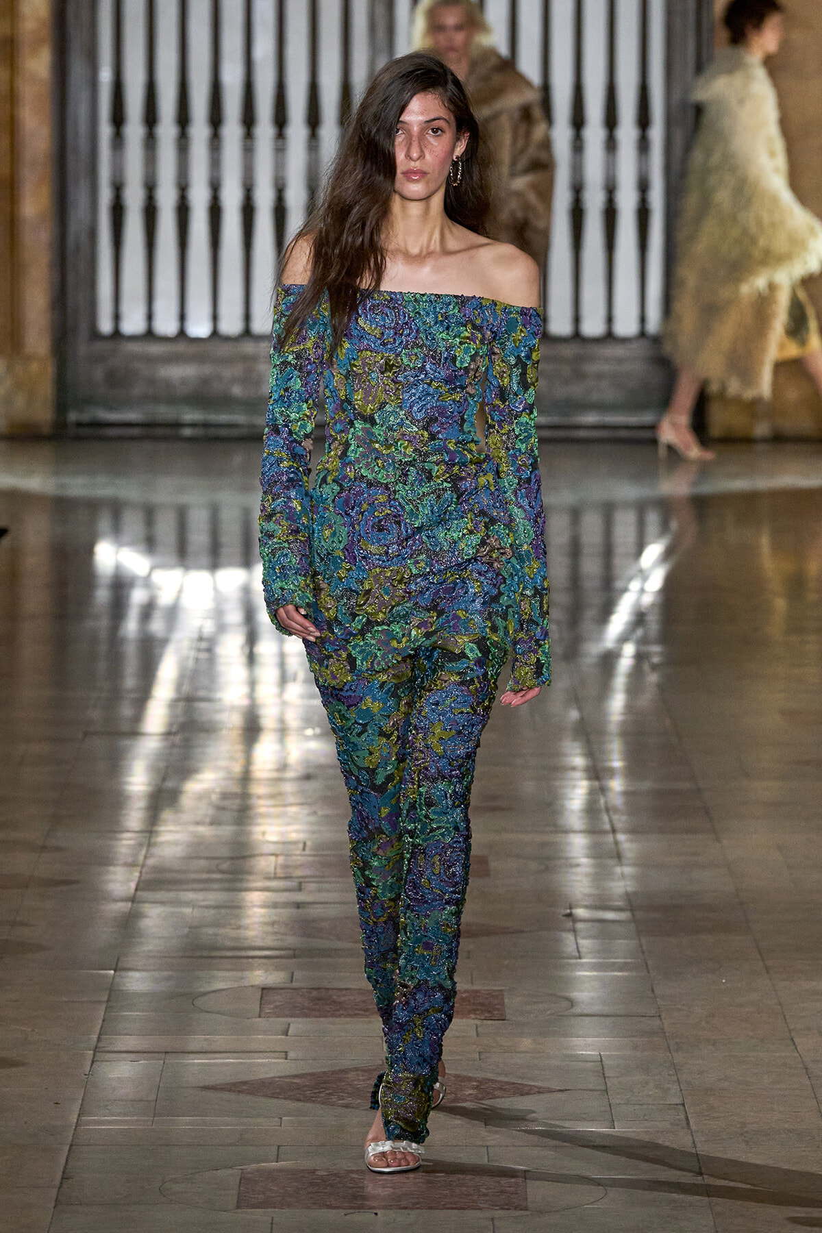 Model on a runway wearing a colorful off-the-shoulder floral jumpsuit with long sleeves and silver sandals.