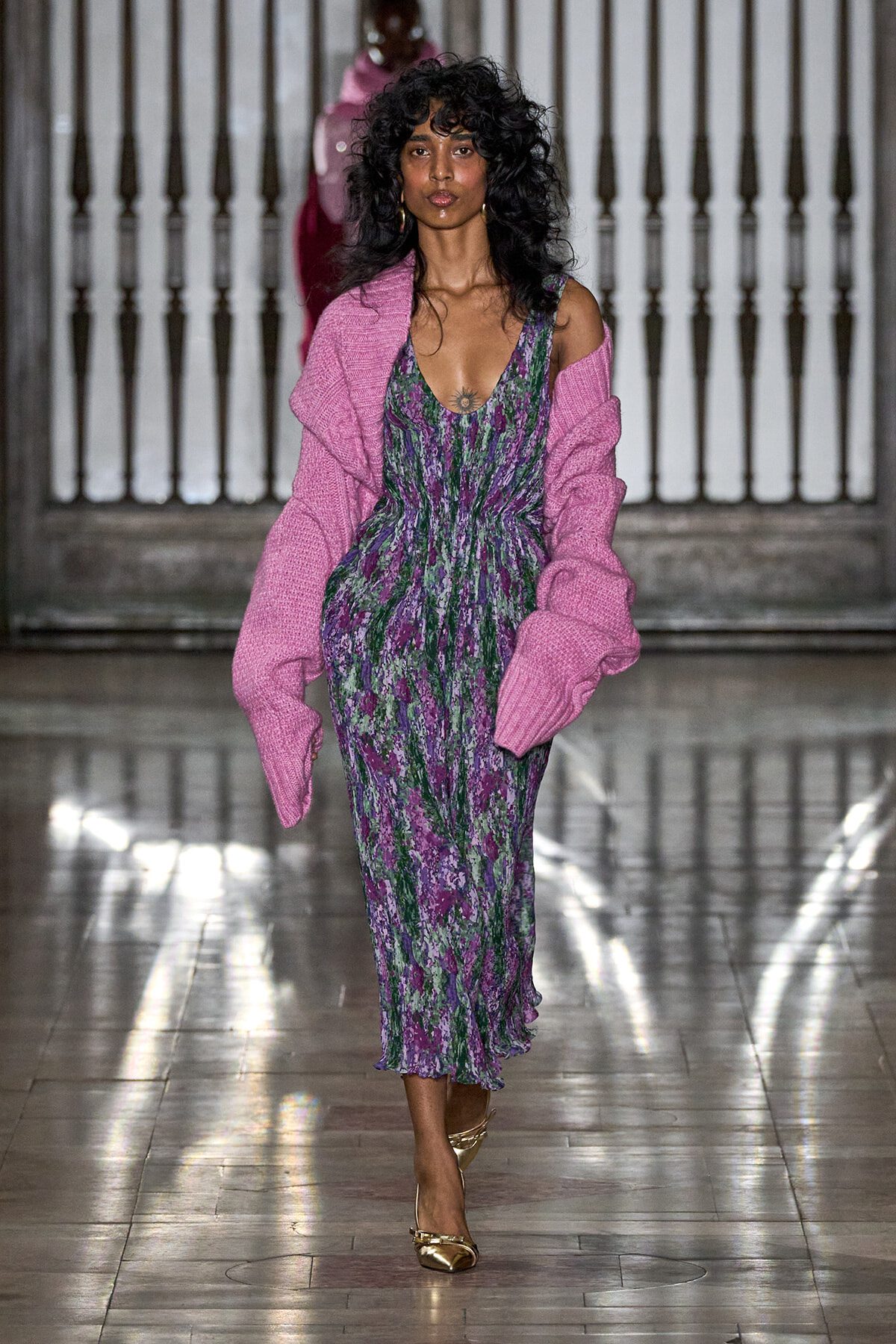 Female model walking a runway in a purple-green floral midi dress with a pink knit cardigan and gold heels.