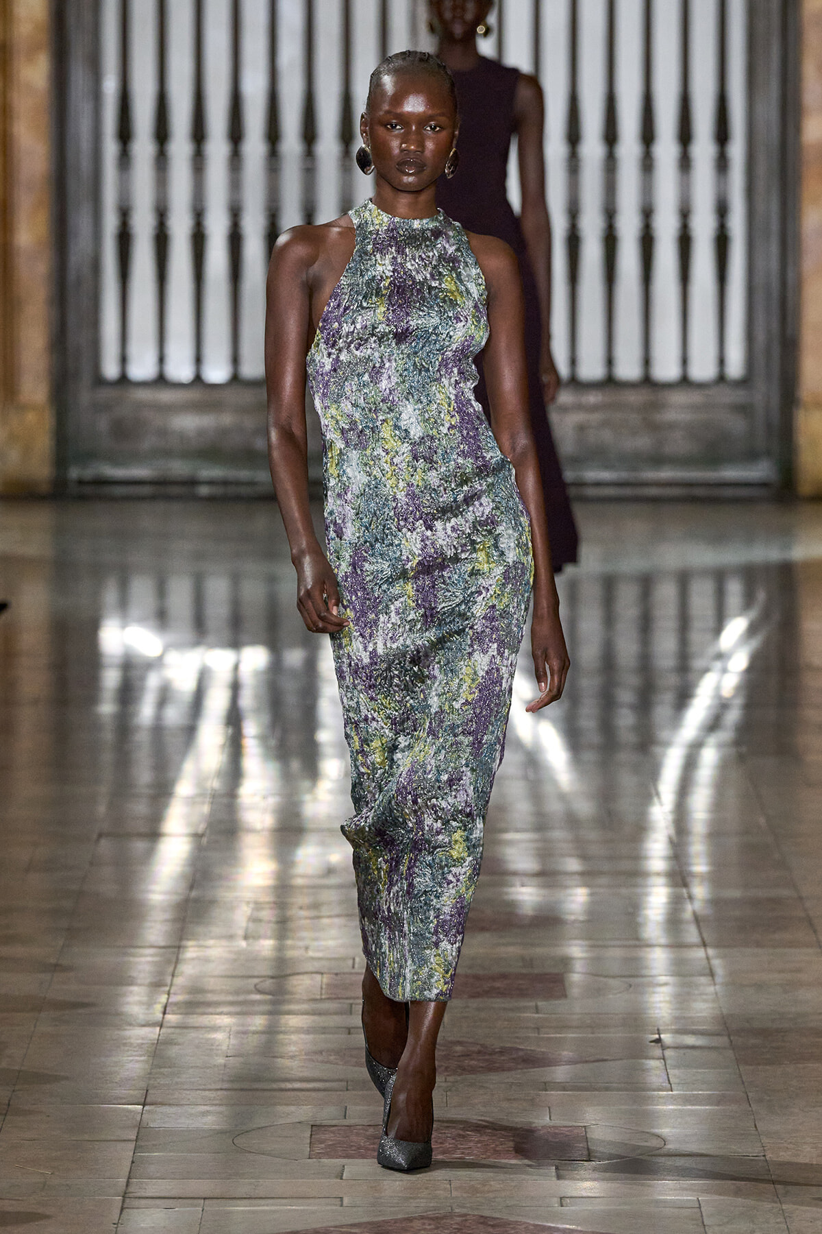 Model walking the runway in a sleeveless high-neck midi dress with a green‑purple floral pattern, wearing large hoop earrings in a historic venue.