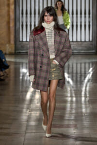 Model walking the runway in an oversized plaid coat over a cream turtleneck and olive distressed mini skirt, white heels.