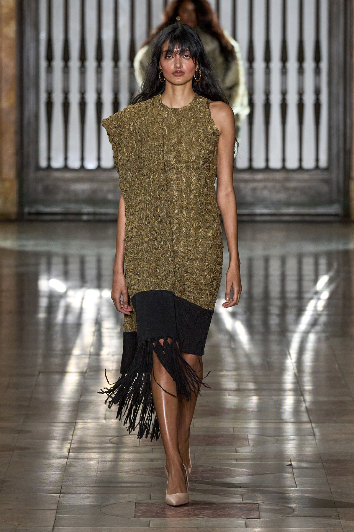 Model walking a fashion runway in a metallic gold textured sleeveless dress with a black fringe hem and beige heels.