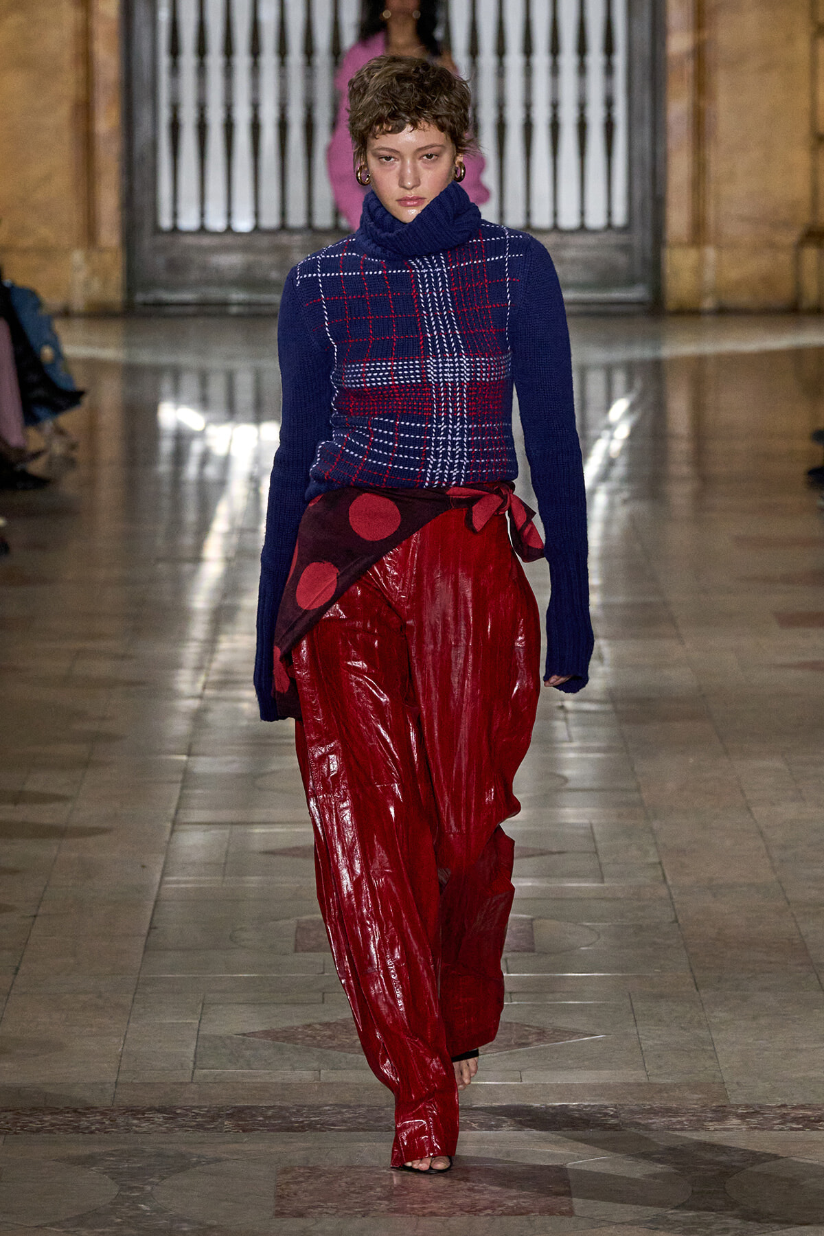 Model on a runway wearing a blue turtleneck sweater with a plaid pattern and glossy red wide-leg pants with a tied waist wrap.
