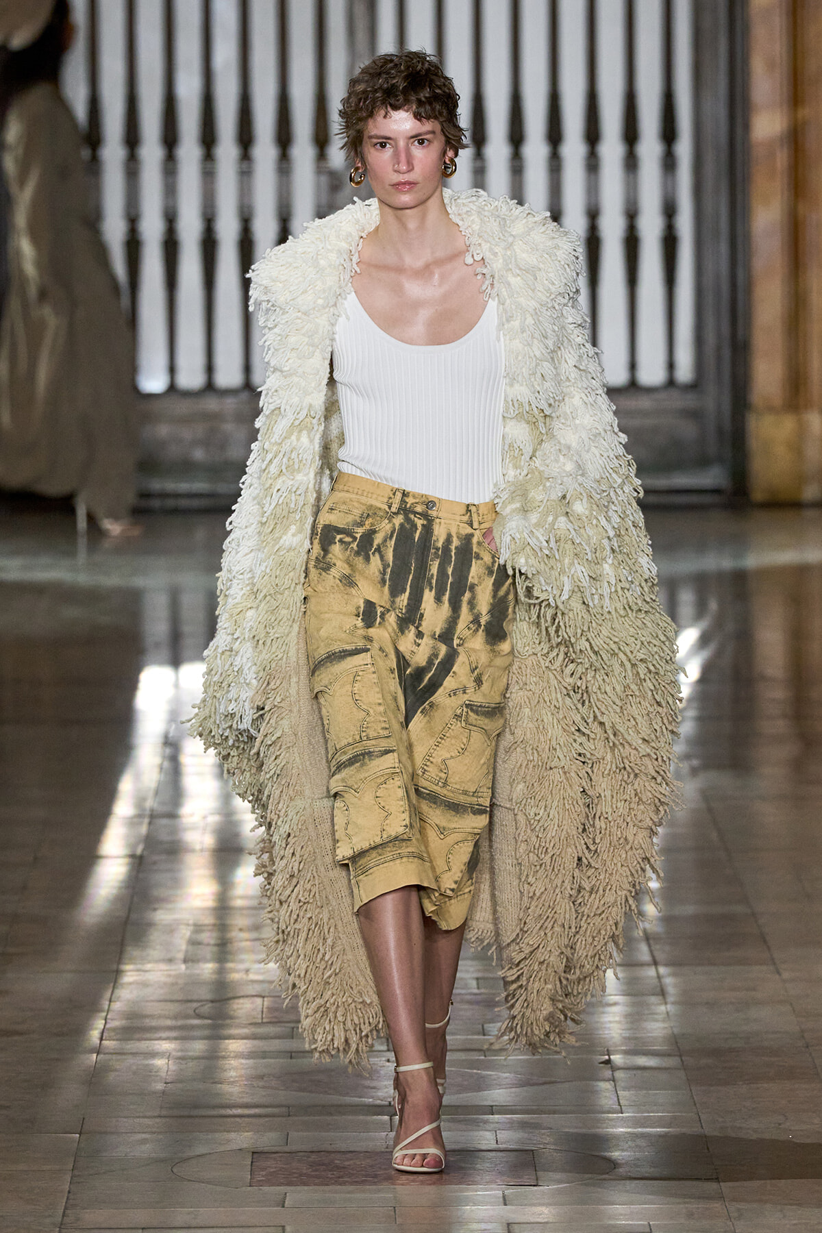 Model walking the runway in a long cream shaggy coat over a white ribbed tank and tan cargo skirt with strappy sandals.