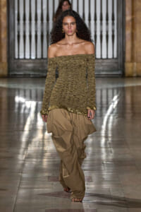 Female model walks a fashion runway in a gold off-the-shoulder knit top and oversized tan pants, with a neutral backdrop.