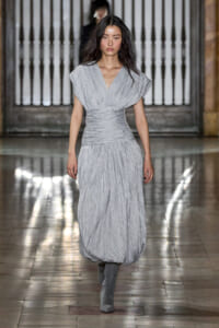 Model on a fashion runway wearing a light gray, pleated wrap dress with a deep V-neck and ruched waist, paired with gray knee-high boots.