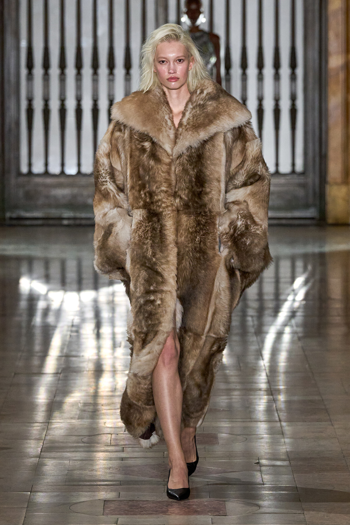 Model walks down a fashion runway wearing a voluminous brown fur coat and black heels, blonde hair, in an indoor showroom setting.