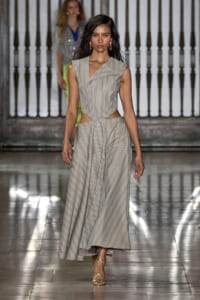 Model walks the runway in a beige-gray striped two-piece dress with waist cutouts and a full, flowing skirt; metallic sandals and gold hoop earrings visible.