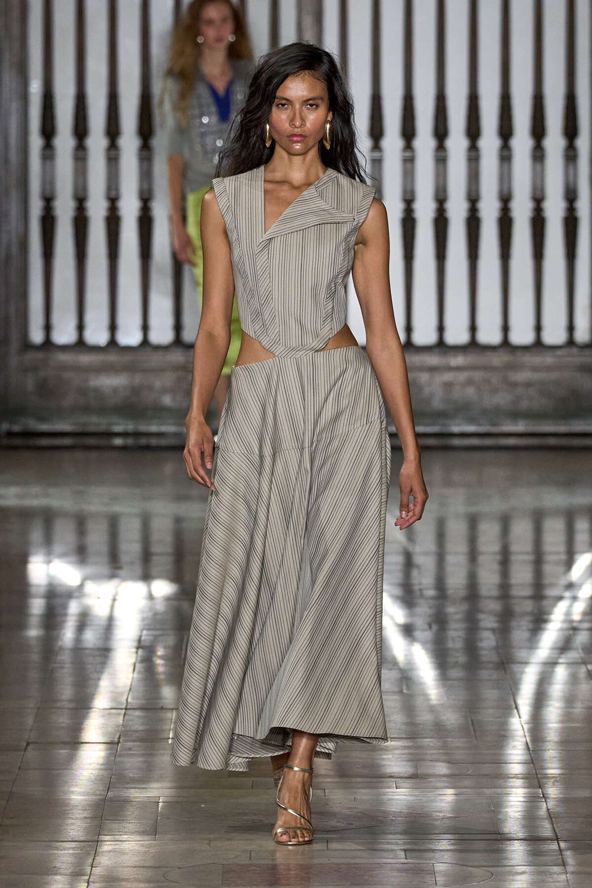 Model walks the runway in a beige-gray striped two-piece dress with waist cutouts and a full, flowing skirt; metallic sandals and gold hoop earrings visible.