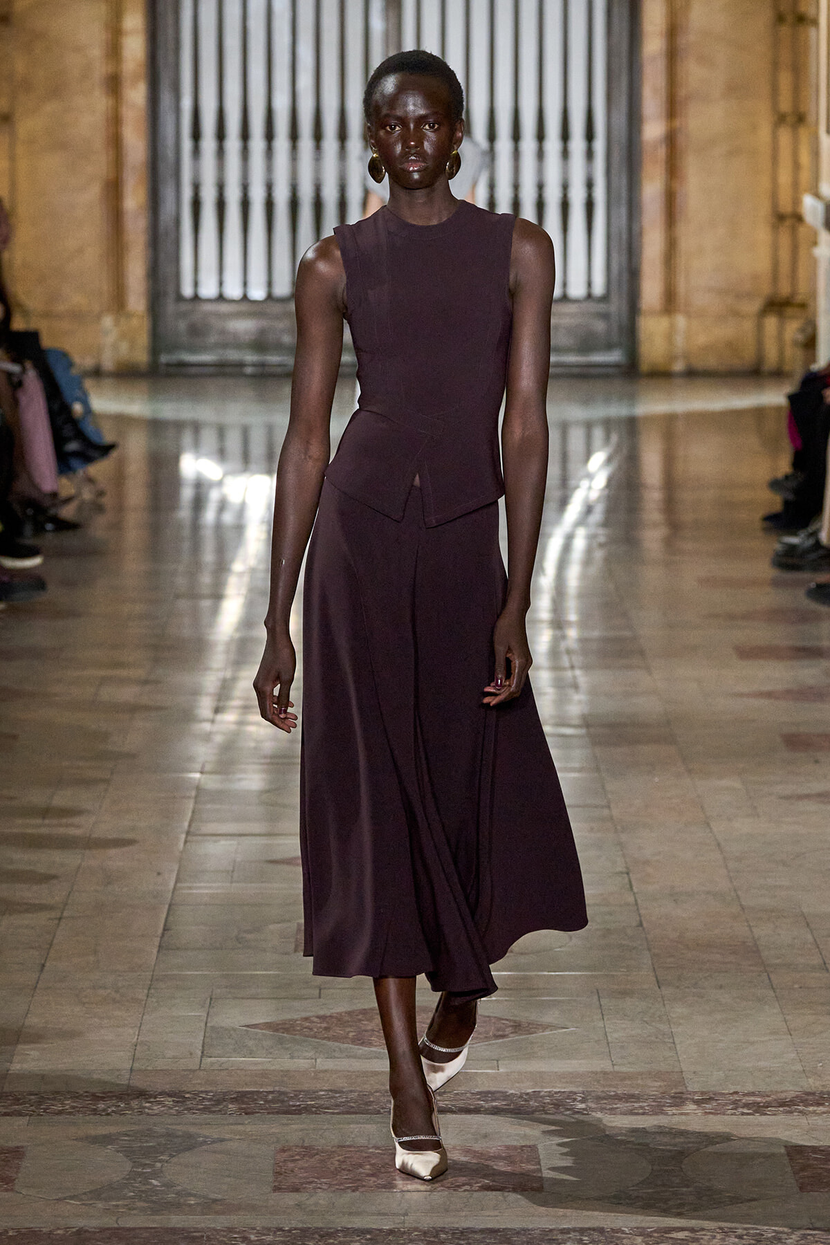 Model walks the runway in a sleeveless burgundy dress with a wrap-front skirt, in a grand hall venue.