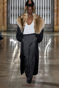 Model walking runway in a white tank, oversized black coat with a large fur stole, and pinstripe trousers.
