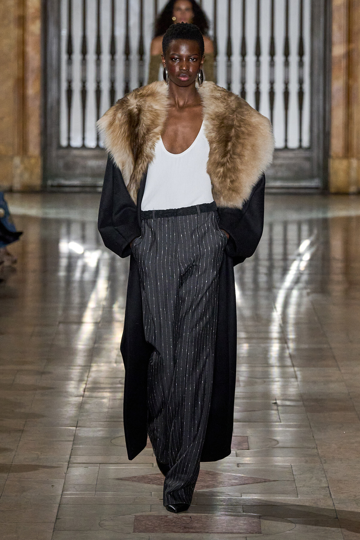 Model walking runway in a white tank, oversized black coat with a large fur stole, and pinstripe trousers.
