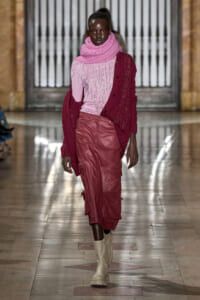 Model walks a runway in a pink top with fringe, a pink scarf, a burgundy cardigan, red cargo skirt, and beige boots in a marble hallway.