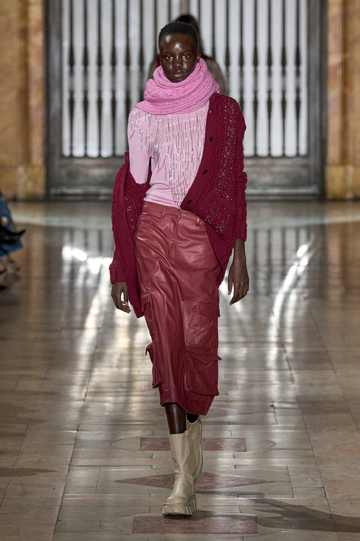 Model walks a runway in a pink top with fringe, a pink scarf, a burgundy cardigan, red cargo skirt, and beige boots in a marble hallway.