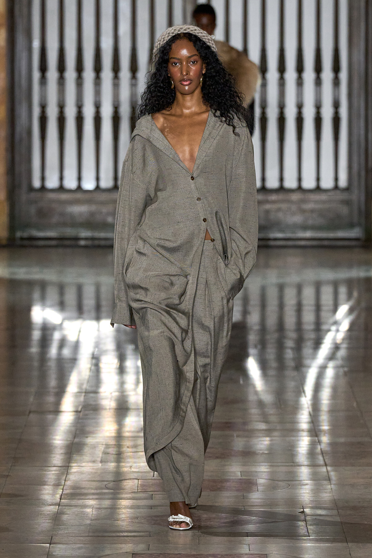 Model walking a runway in an oversized gray suit with a deep V-neck and headband, on a reflective tiled floor.