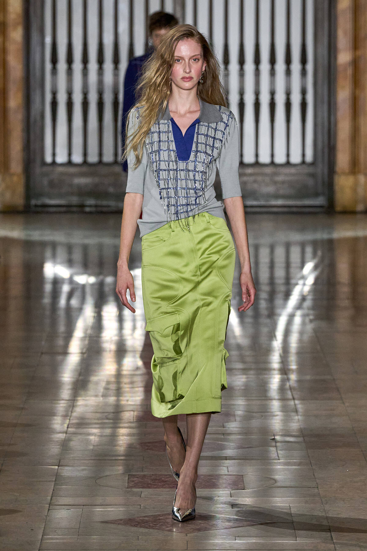 Model walks the runway in a gray polo-style top with blue V-neck and grid embroidery, paired with a lime-green satin midi skirt.
