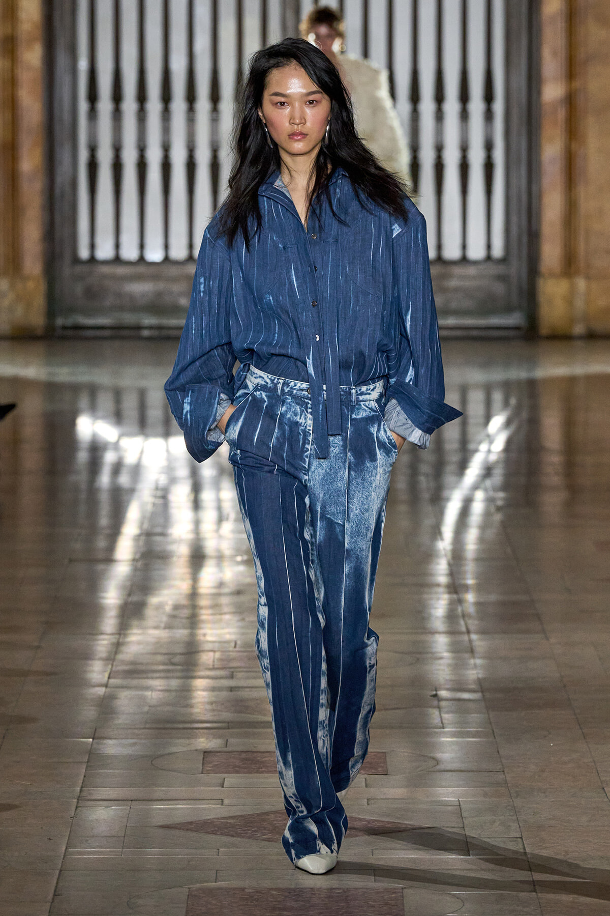 Model walking a runway in a blue denim outfit with vertical white stripes; matching loose shirt and wide-leg jeans with tie-dye accents, hands in pockets.