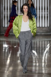 Model walking a fashion runway in a lime-green oversized jacket with a fur collar, white top, and gray pinstriped wide-leg trousers.