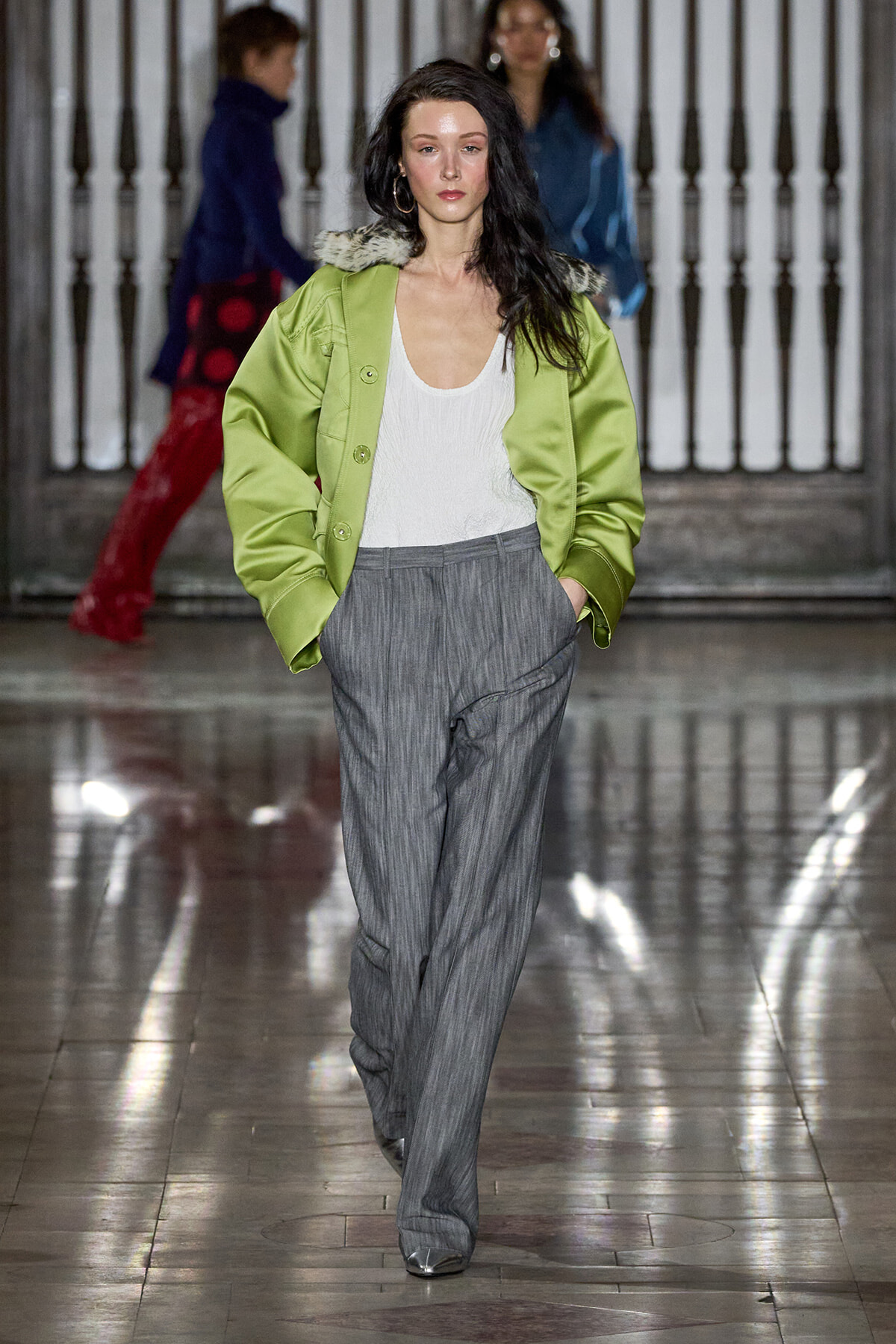 Model walking a fashion runway in a lime-green oversized jacket with a fur collar, white top, and gray pinstriped wide-leg trousers.