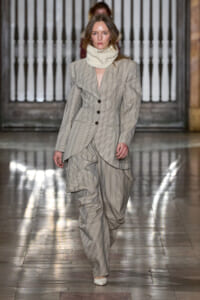 Runway look: a model wears a light pinstripe suit with a layered, draped front and oversized trousers, paired with a chunky cream knit scarf.