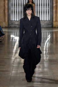 Model walks a fashion show wearing a tailored black blazer and wide, draped black pants on a marble runway