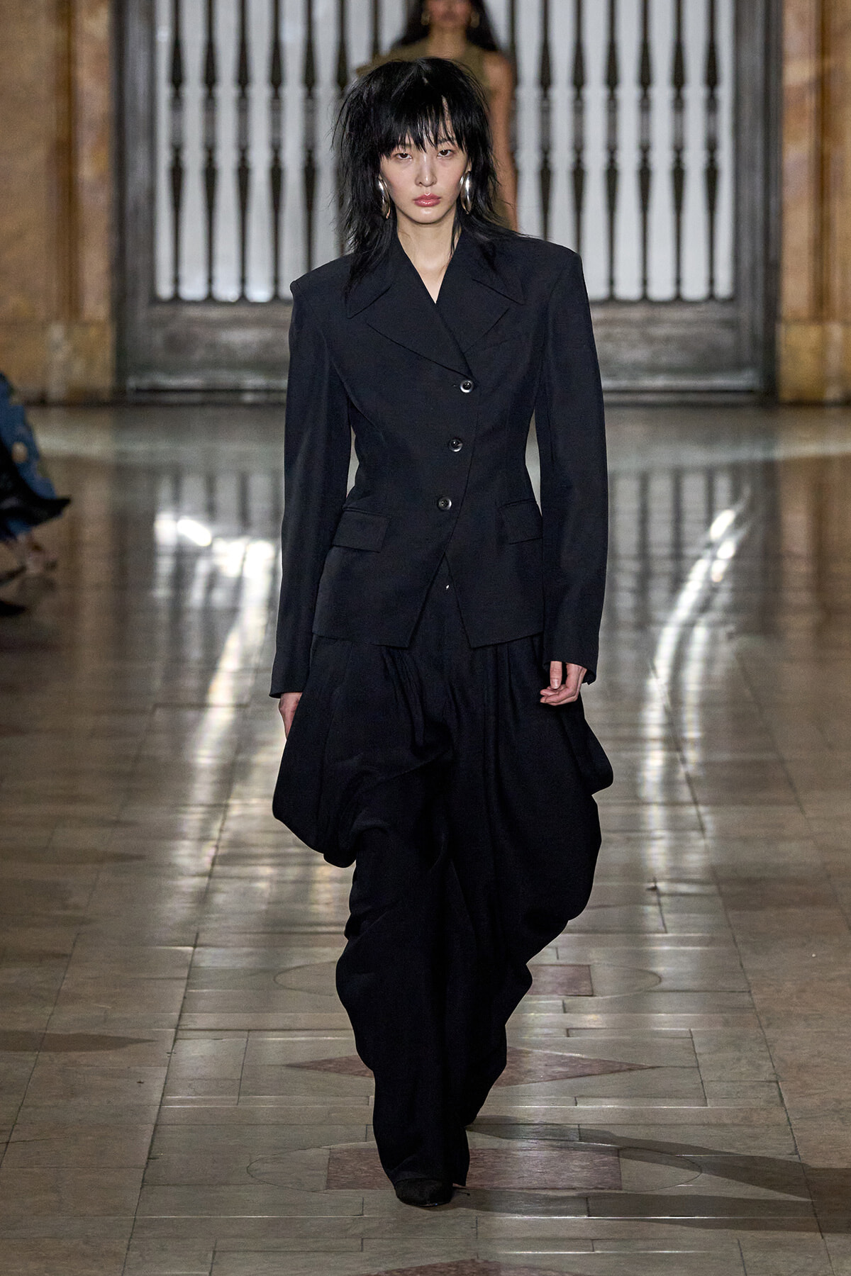 Model walks a fashion show wearing a tailored black blazer and wide, draped black pants on a marble runway