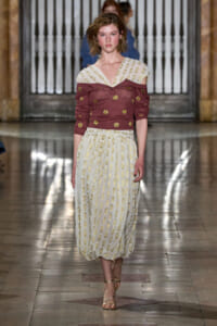 Model walks a runway in a burgundy off-shoulder knit with gold dots and a cream, gold-dotted pleated skirt.