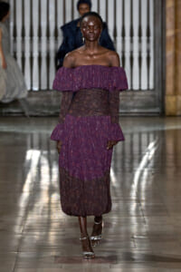 Female model walking a runway in a purple off-the-shoulder gown with a sheer waist and silver sandals.