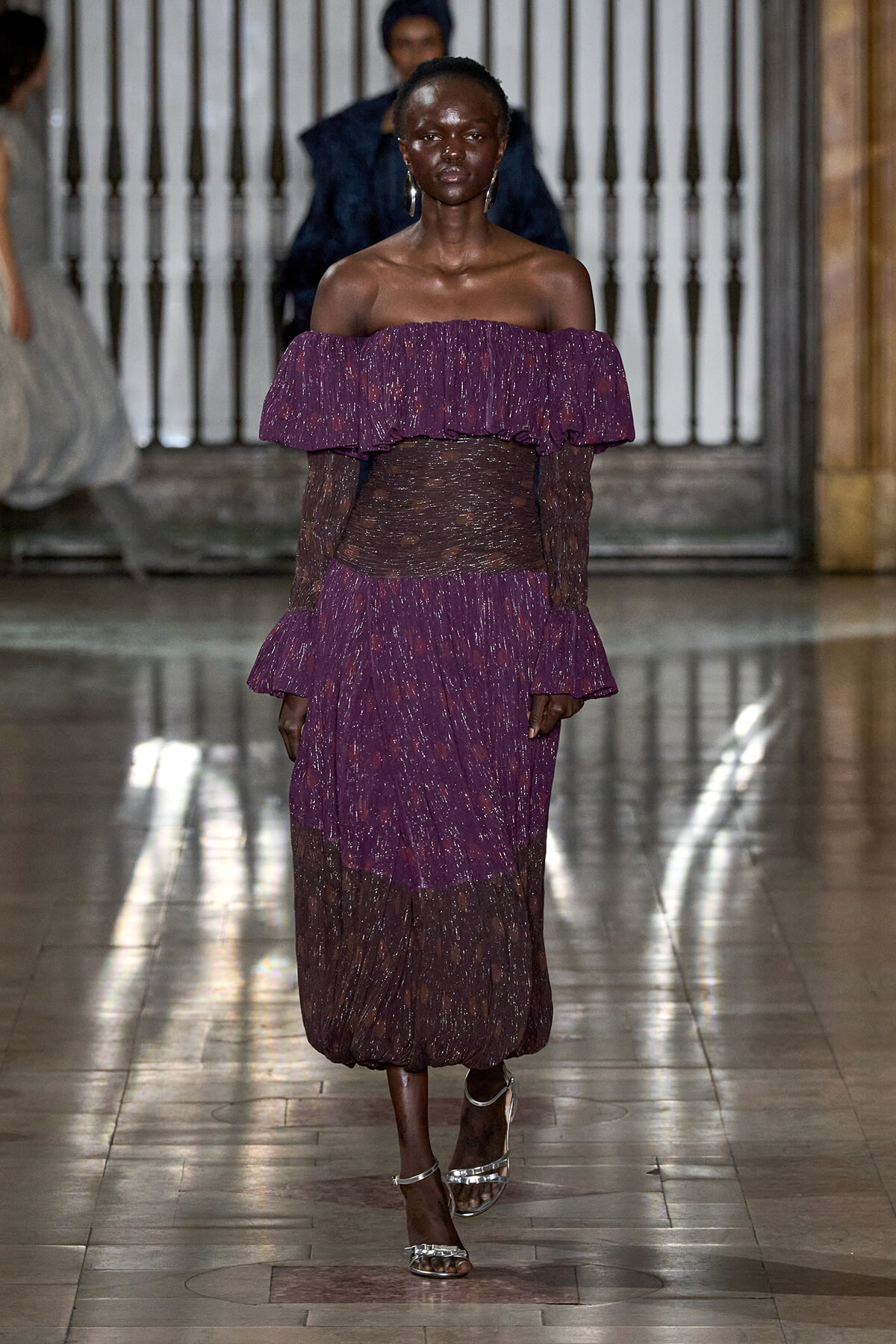 Female model walking a runway in a purple off-the-shoulder gown with a sheer waist and silver sandals.