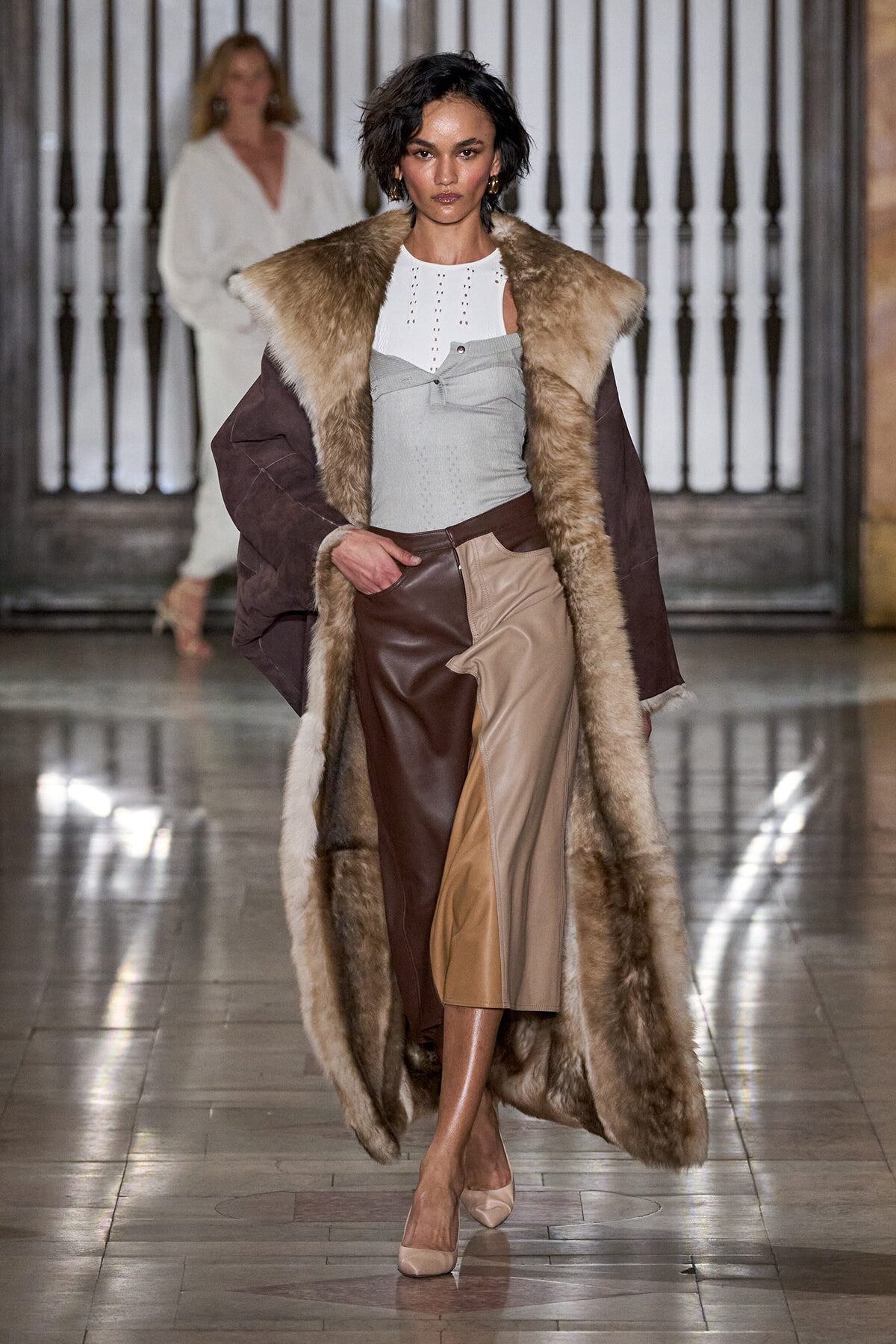 Model walking a fashion runway in a long brown fur coat over a white top and color-blocked leather pants, beige heels.