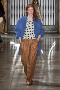 Model walking runway in a blue oversized coat over a cream polka-dot blouse and brown wide-leg trousers, hands in pockets.